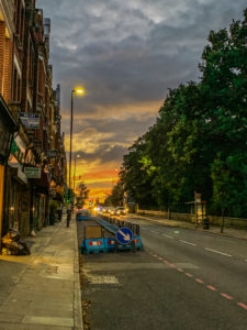 colourful sunset set in warm oranges and yellows along a road, lined with lush trees on one side and brick buildings on the other side