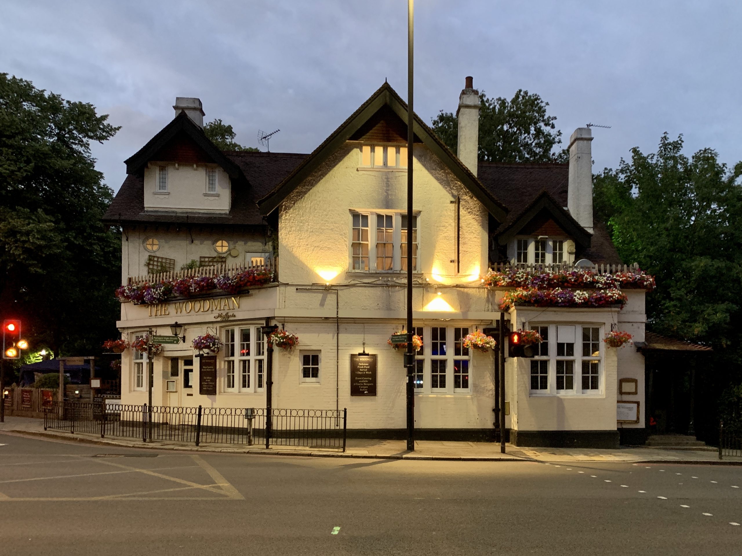 white two-storey building behind a street crossing, with a gabled roof, lit up by spotlights and a lamp post, during dusk