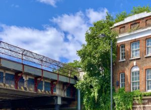 sky with rusting railway bridge and barbed wire on its fence, next to it, a half overgrown brick house. sunny day.