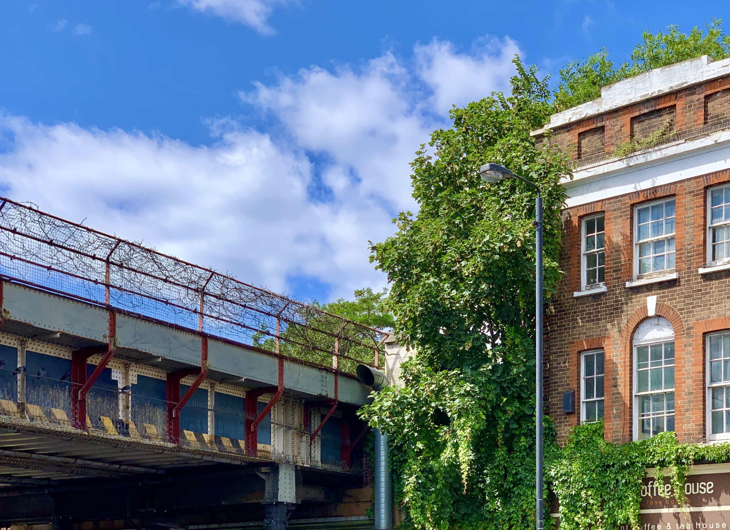 sky with rusting railway bridge and barbed wire on its fence, next to it, a half overgrown brick house. sunny day.