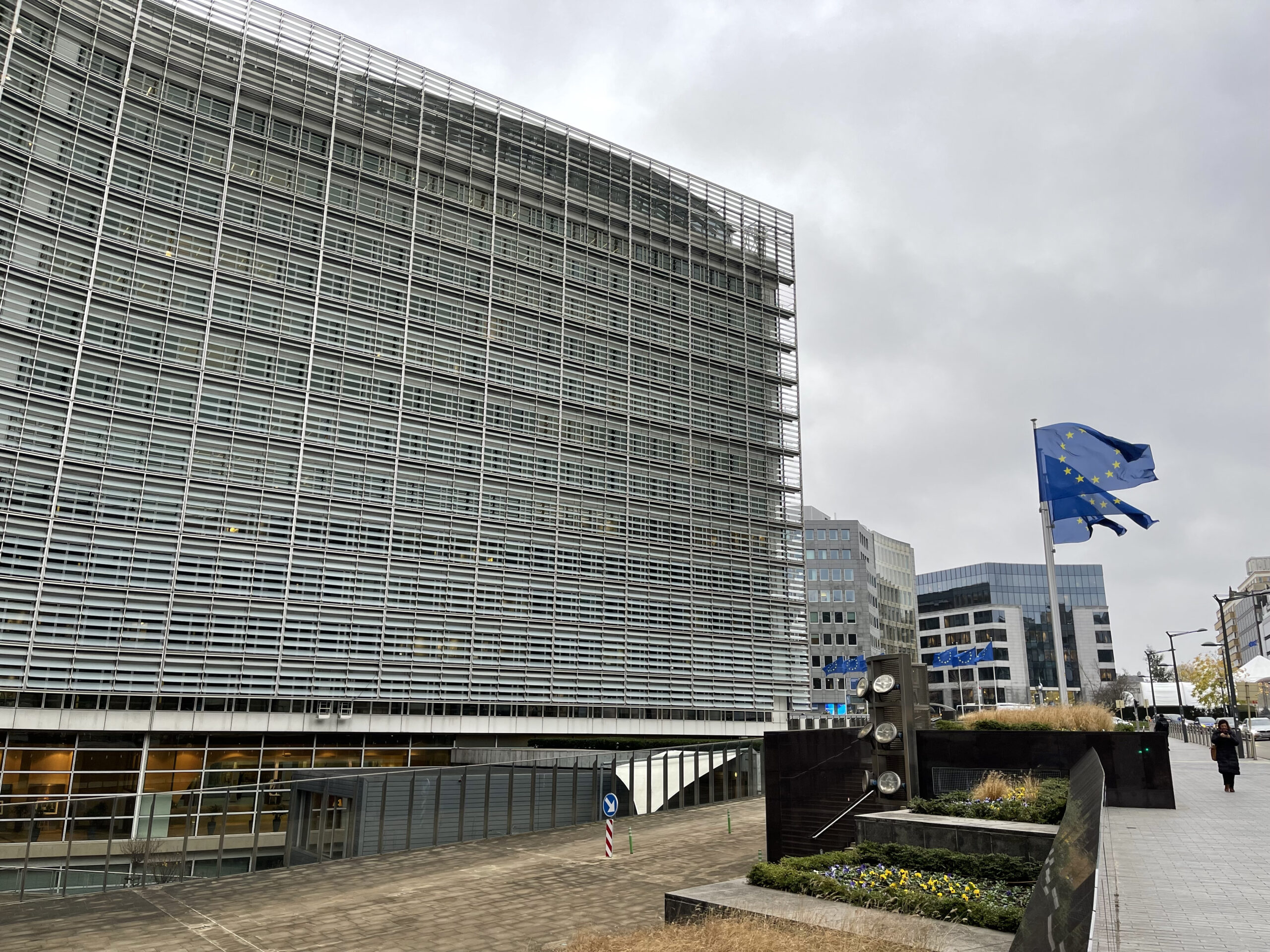 The curved Berlaymont building in Brussels to the left, and 3 EU flags blown by the wind on the right. The sky is grey in the background.